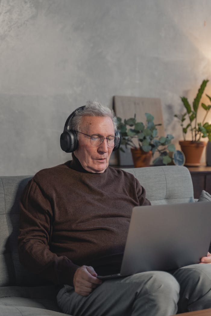 Elderly man using laptop with headphones for online meeting at home, enhancing digital communication skills.